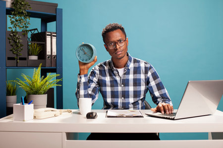 Project team leader sitting at desk with a clock in his hand, showing annoyance at his employees...