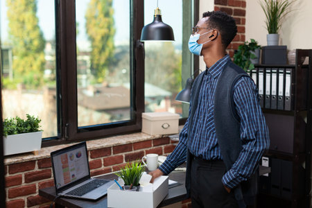 Startup employee wearing covid mask looking out the window with regret after being fired from work....