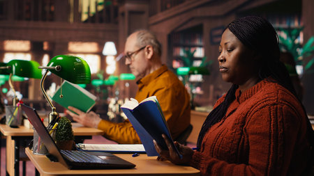 African american woman with a book in hand studying in a quiet library space, working hard to...