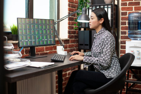 Asian female entrepreneur researching financial trends on computer in brick wall room. Businesswoman...