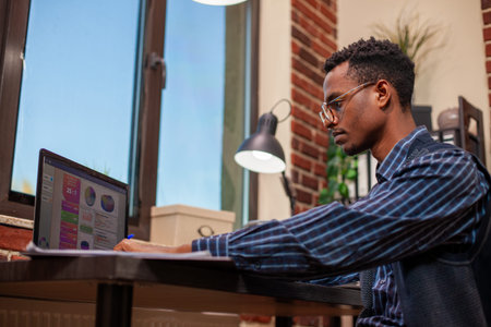 African American manager at office desk reviewing company research updates on his laptop. Male...