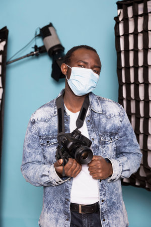 Portrait of a black cameraman protecting himself with a face mask while standing in blue background...