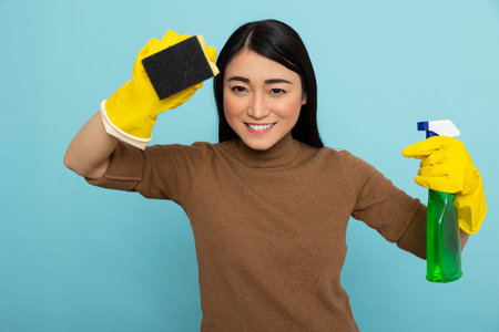 Cheerful housemaid in rubber gloves posing with cleaning supplies, showing positive attitude in...