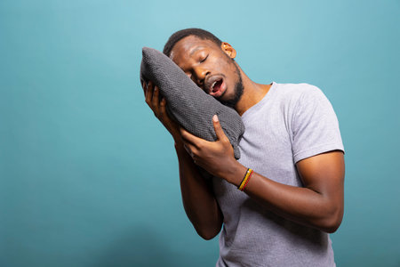 Tired young man with mouth open and eyes closed, resting his head on a pillow, showing exhaustion....