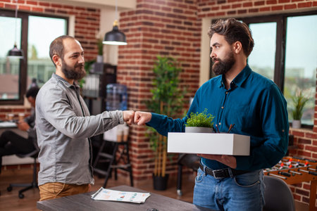 Departing male employee holds a box of belongings and exchanges a fist bump with a colleague, in...