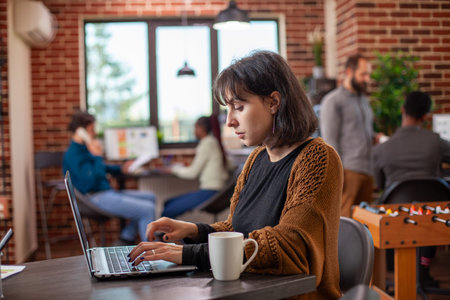 Caucasian businesswoman seated at desk, using laptop in modern office with coffee cup nearby. Young...