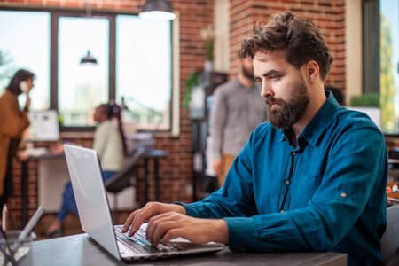 Bearded businessman seated at office desk, typing on laptop in modern workspace. Caucasian male...