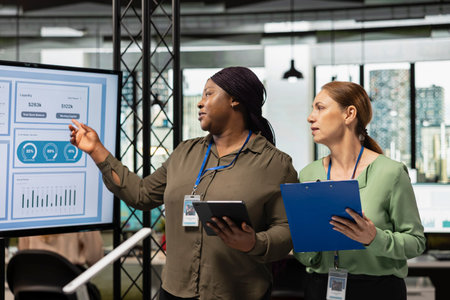 Diverse businesswomen gather around documents and TV monitor as a unified team to work on planning,...
