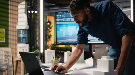 Middle eastern real estate developer evaluates construction site blueprints on laptop, working on...