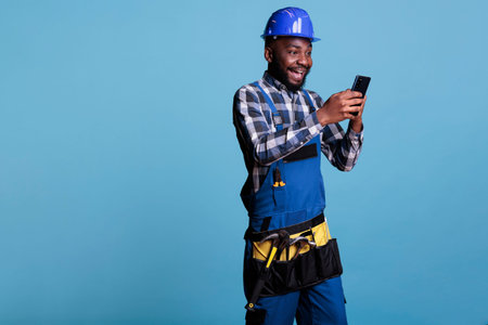 African american builder receiving good news via text message on cell phone. Optimistic contractor wearing work uniform, hard hat against light blue background in studio shot.の写真素材