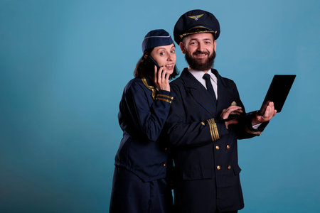 Smiling pilot using laptop, flight attendant talking on smartphone. Airlane captain in professional...