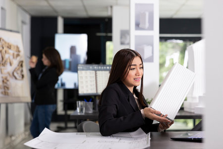 Asian woman reviews architectural prototype during video call, carefully analyzing project details with team. Female architect holds 3d model, planning new buildings for construction with remote clientの写真素材