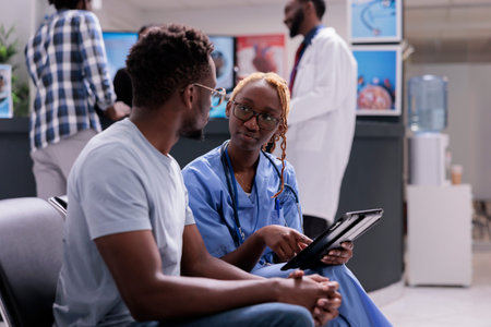 African american nurse and patient talking about disease, doing checkup consultation with sick man in hospital waiting area. Medical assistant examining person to give medication, health center.の写真素材