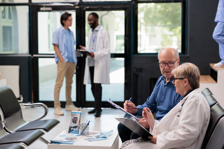 Senior doctor sitting on chair in hospital reception explaining disease diagnosis to elderly patient...