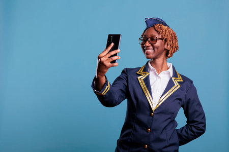Smiling female flight attendant wearing uniform having video call with mobile phone. African...