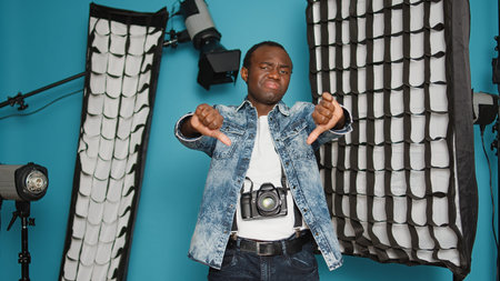 Young man showing thumbs down dislike gesture in studio with professional photography equipment. Expressing disagreement and disapproval, having tripod, and lighting backstage.の写真素材