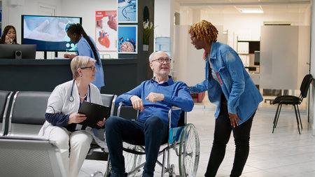Caretaker giving social assistance to old wheelchair user at consultation appointment with physician. Senior person with chronic disability attending checkup visit with medic and carer.の写真素材