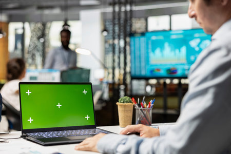 Caucasian businessman seated in office, looking at device with green screen display. White male employee works on laptop with isolated copy space template, focusing on financial strategies.の写真素材