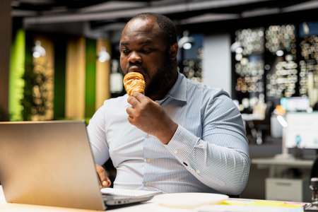 Black person dealing with a stress eating habit in office after hours, overeating and being under pressure from intense workload. Living an unhealthy lifestyle with negative emotions at work.の写真素材