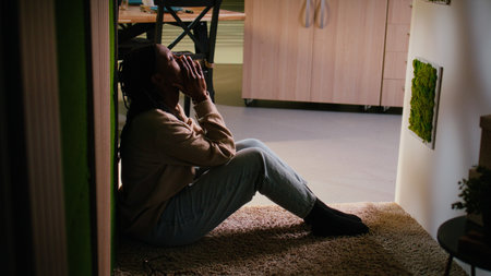 Exhausted worried black woman struggling with depression and stress, sitting alone on the floor,...
