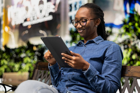 Happy woman using tablet, sitting on city bench, relaxing by looking at social media posts on feed. Cheerful young girl in park uses digital device to browse online content, urban landscape backgroundの写真素材