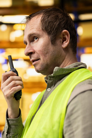 Professional warehouse employee using wireless radio transmitter to coordinate cargo distribution. Tracking technology and walkie-talkie signal support organized e-commerce operations.