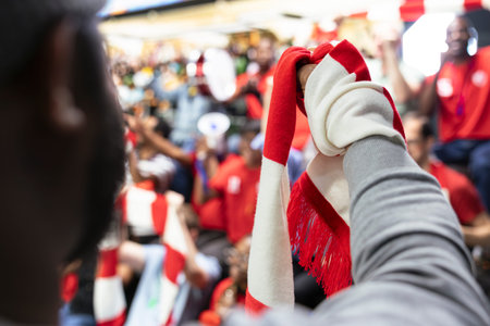 Close-up of supporter raising a red and white scarf during live soccer match, with chanting stadium crowd and fan atmosphere behind. Diverse multigenerational sports fans join the chant.