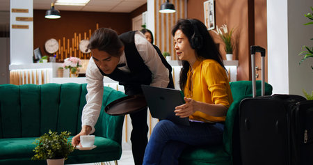Asian woman on laptop ordering coffee in lounge area, watching tv show on PC while she waits for her room to be available. Staff serving drink from hotel bar, tourist receiving great services.