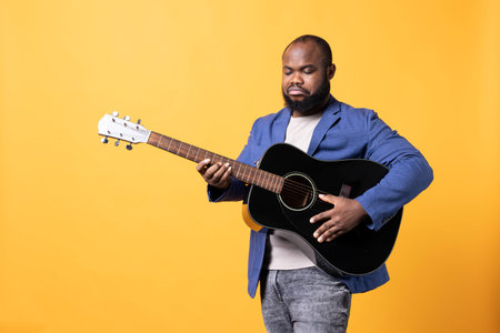 BIPOC man tuning guitar before performing songs during concert, isolated over studio background. Upset artist unhappy with out of tune stringed acoustic musical instrument, fixing it.