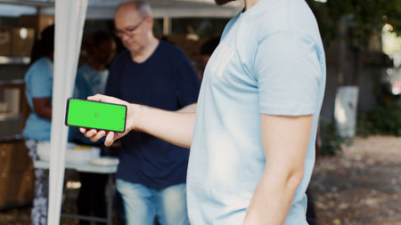 Person horizontally holding mobile phone at charitable food drive, presenting an isolated mockup template, volunteer grasping smartphone with blank green screen display, tripod shot.