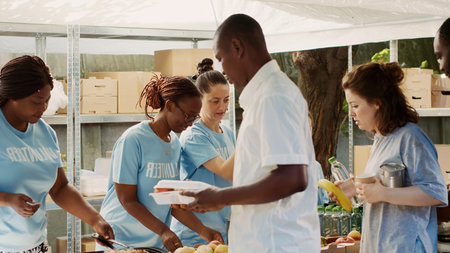 Multiracial volunteer ladies serve meals and non-perishable products to hungry and homeless people, friendly charity workers handing out free food to the poor and needy.