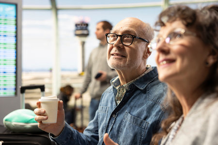 Smiling elderly man and woman, drinking coffee, in airport lounge area, discussing ideas and travel plans before flight. Confident retirees representing active lifestyle and fulfilling life goals.