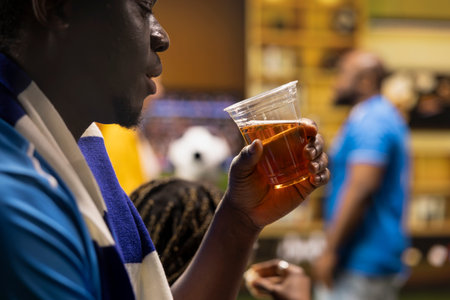 Close up of football supporter drinking beer from plastic cup while live match plays on TV in living room. Side view of man enjoying alcohol beverage during relaxed home watch party
