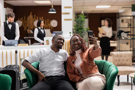 Black couple smiling for pictures and resting on lounge area couch, luxury hotel reception lobby. Travelers waiting to check-in and start their vacation, excellent guest service and hospitality.