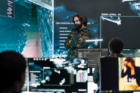 Military analyst evaluates enemy tactical activity on surveillance feed projected on large display. Intelligence officer checks digital maps, doing geospatial analysis