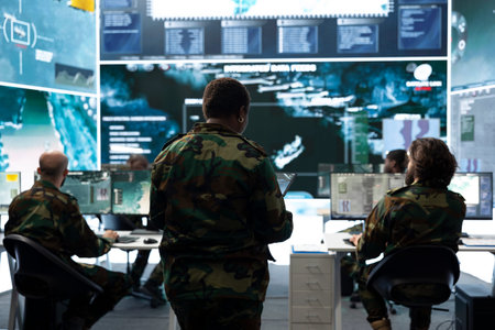 Team of military officers monitoring surveillance displays across operations room. Army supervisors overseeing operators tracking satellite maps and target information