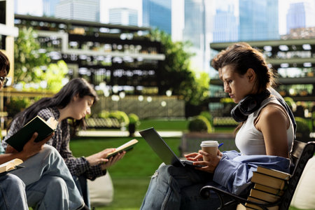 Focused student working on laptop and holding coffee cup on campus bench while classmates reading nearby. Young woman with headphones behind neck, collaborative learning.