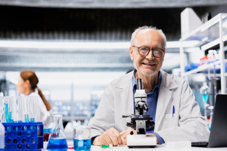 Portrait of smiling senior pharmaceutical scientist inspecting new drug compound under microscope for consistency. Jolly elderly laboratory employee verifying medication quality and purity standards