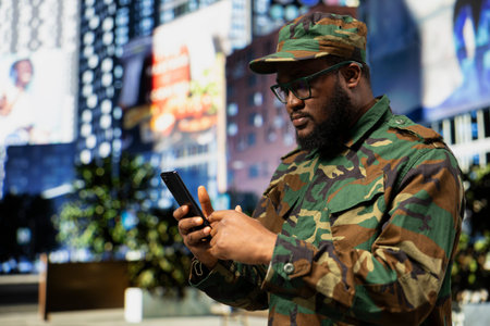 Confident soldier in tactical uniform checking mobile phone screen, navigating web page with disciplined focus as he stands on a city street, representing commitment for the military.