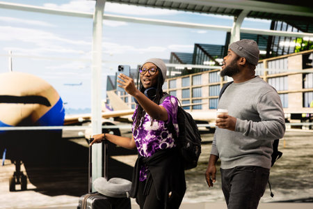 Two travelers taking smartphone selfie in modern airport terminal, carrying trolleys and backpacks. Friends walking in airport in front of plane behind windows glass, taking photos with phone camera