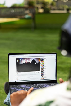 Black young adult looking at live concert performance on streaming platform, laptop running digital media content for music entertainment. Study break on campus.