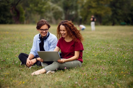 Profile of a two smiling colleagues, seated on a green grass, looking on a laptop, isolated on a...