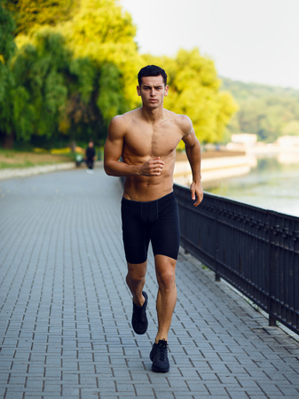 A frontal pictures of a young athletic, fit, muscular man shirtless, running morining in park, isolated on nature background. Workout lifestyle.Vertical view.の写真素材