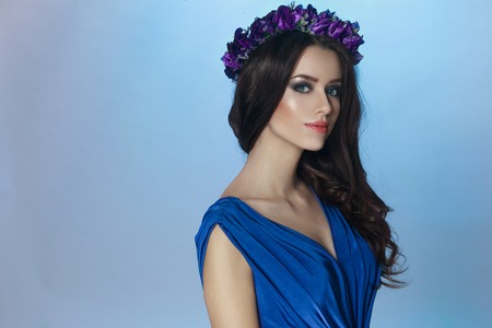 A beautiful fashion brunette model with splendid make up and curly long hair and crown with violets flowers on her head, in blue dress, looking at camera. Horizontal portrait on the blue background.の写真素材