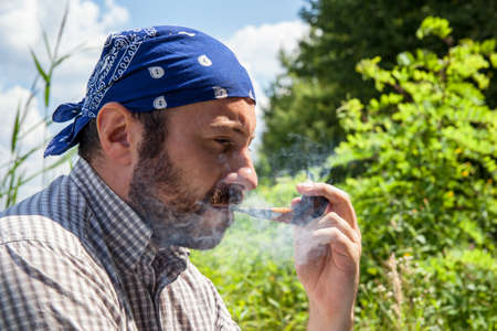 Bearded man smoking his pipe and releasing smoke outside on a summer dayの写真素材