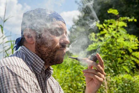 Bearded man enjoying his pipe and releasing smoke outside on a summer dayの写真素材