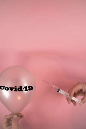 Hands holding vaccine syringe with needle and balloon with Covid-19 writing. On soft pink background. Minimal abstract corona virus  pandemic concept. Stay safe.の写真素材