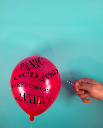 Red balloon with panic, anxiety, depression, PTSD and OCD writings. With hand holding a needle. On soft aquamarine background. Minimal abstract disorders concept.の写真素材