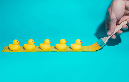 Five rubber ducklings in a row on yellow paint, following hand with brush, on trendy blue background. Minimal abstract summer, teamwork or fashion concept. With copy space.の写真素材