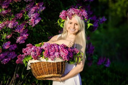 Beautiful smiling young woman is wearing wreath and basket of lilac flowers, white dress, sunsetの写真素材